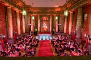 Elegant ballroom filled with guests at round tables, featuring red decor and a checkered dance floor under ambient lighting.