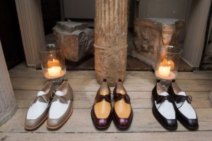 Two pairs of stylish shoes with contrasting colors placed near candles and ancient-style columns on a wooden floor.