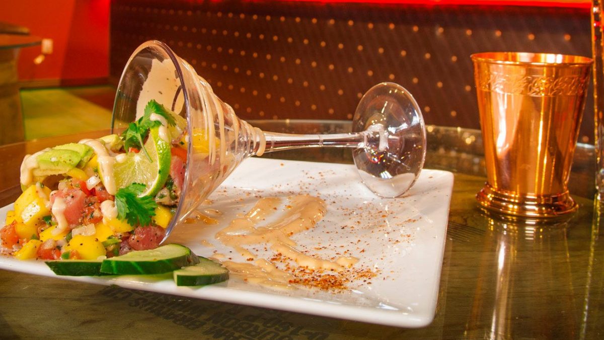 A spilled fruit salad with mango, cucumber, and cilantro, served in a glass, on a white plate with sauce drizzled around.