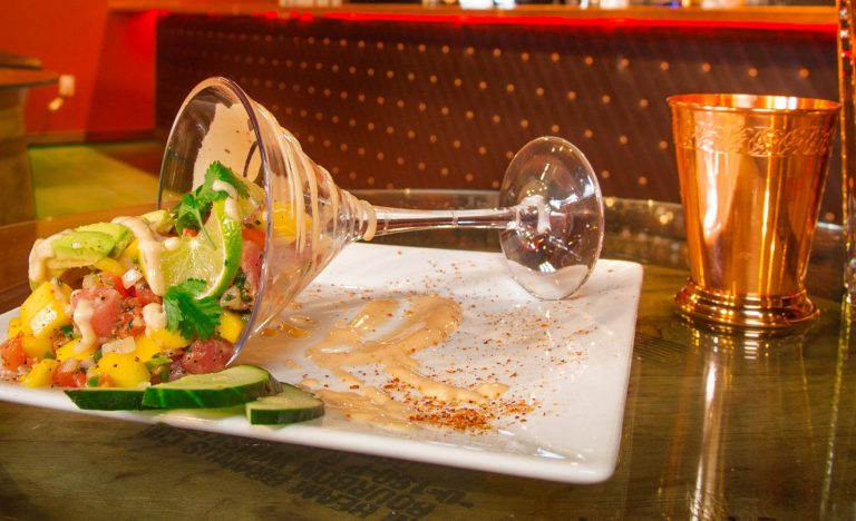 A spilled fruit salad with mango, cucumber, and cilantro, served in a glass, on a white plate with sauce drizzled around.