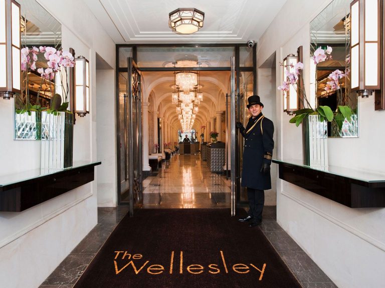 A doorman in a top hat welcomes guests to The Wellesley, featuring elegant decor and floral arrangements in the entranceway.