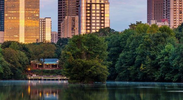 City skyline reflected in a calm lake, surrounded by lush greenery and trees at sunset.