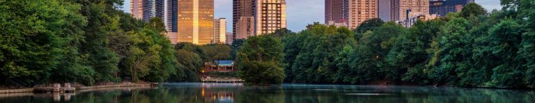 City skyline reflected in a calm lake, surrounded by lush greenery and trees at sunset.