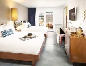 Modern hotel room with two beds, a desk, and a balcony access, featuring stylish decor and a warm color palette.