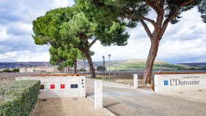 Entrance to Abadia Retuerta LeDomaine with vineyards and hills in the background, framed by towering trees under a cloudy sky.