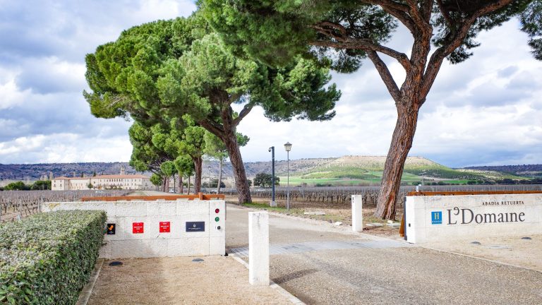 Entrance to Abadia Retuerta LeDomaine with vineyards and hills in the background, framed by towering trees under a cloudy sky.