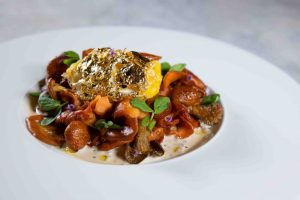 Gourmet dish featuring mushrooms, roasted vegetables, and a golden egg on a creamy sauce, garnished with fresh herbs.