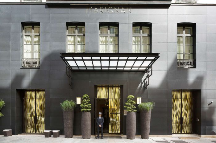 A hotel entrance featuring modern architecture, a doorman in a uniform, and decorative planters on either side.