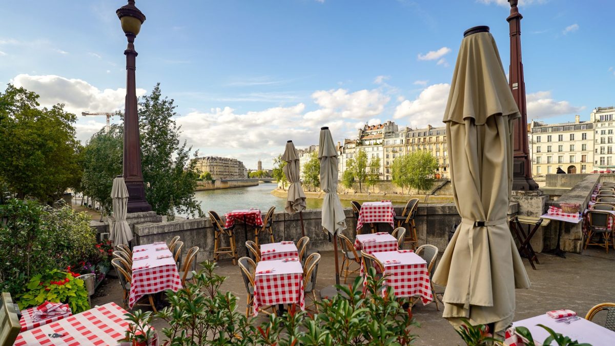 Dining area by the river in Paris featuring tables with red checkered tablecloths and umbrellas, surrounded by greenery.