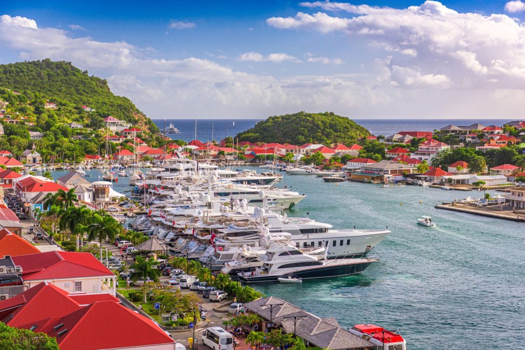 Harbor view of luxury yachts and vibrant red-roofed buildings against a lush green hillside in St. Barth's.