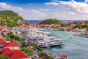 Harbor view of luxury yachts and vibrant red-roofed buildings against a lush green hillside in St. Barth's.