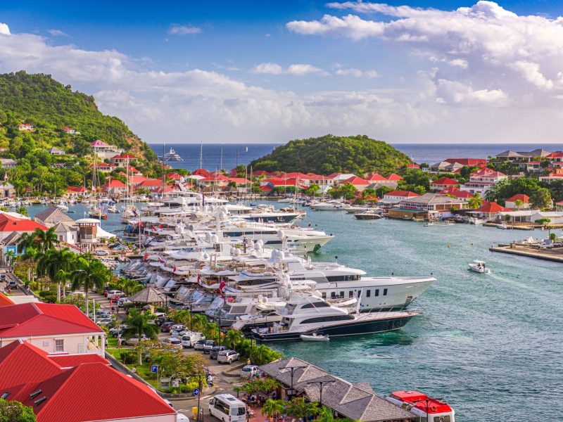 Harbor view of luxury yachts and vibrant red-roofed buildings against a lush green hillside in St. Barth's.