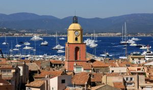 Clock tower overlooking St. Tropez harbor filled with yachts, set against a backdrop of mountains and clear blue sky.