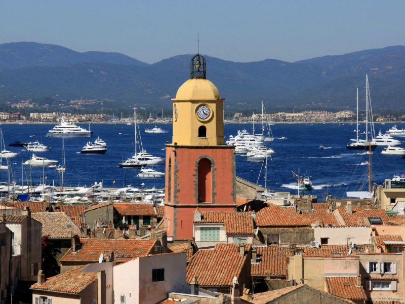 Clock tower overlooking St. Tropez harbor filled with yachts, set against a backdrop of mountains and clear blue sky.