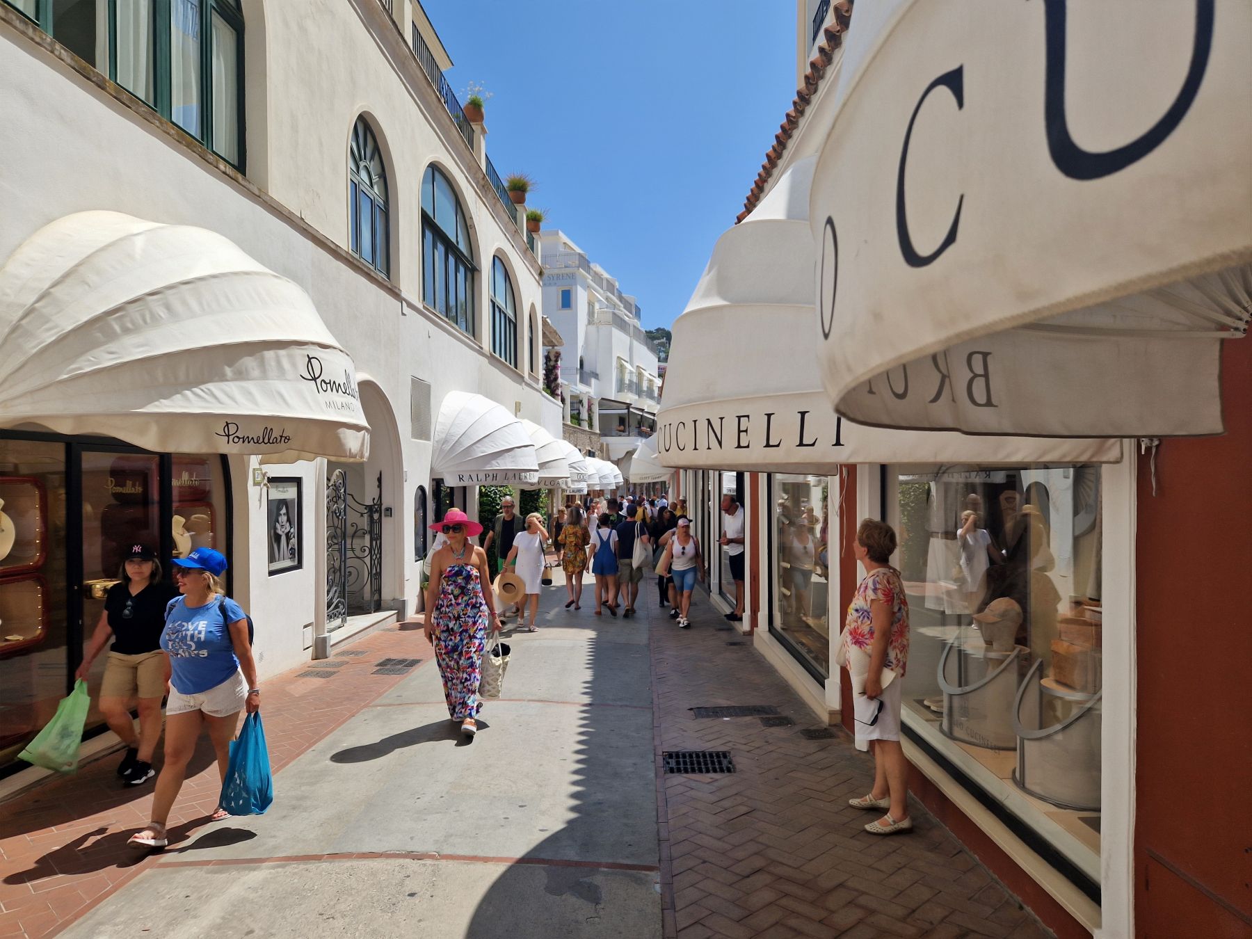 Shoppers stroll through a stylish street in Capri, surrounded by boutique storefronts and sunlit awnings.