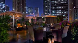 Rooftop lounge at night featuring cozy seating, planters, and city skyline views with glowing lights and umbrellas.