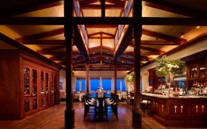 Elegant restaurant interior featuring a bar, wooden beams, and a wine cellar, with tables set against a backdrop of large windows.