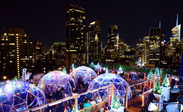 Rooftop setting at night featuring illuminated igloos and a city skyline adorned with festive lights and decorations.