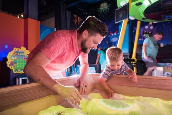 A man and a young boy engage with a colorful sand table in a playful, interactive environment.