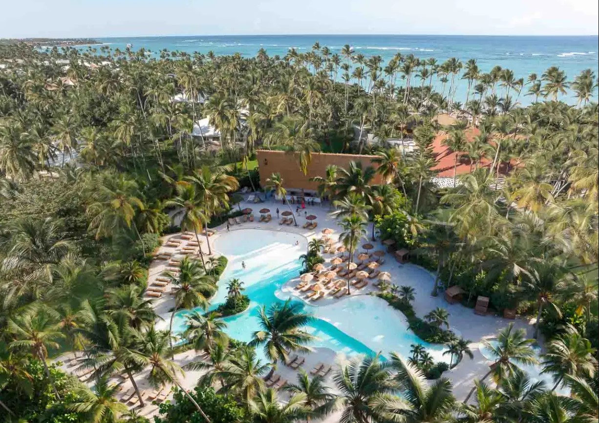 Aerial view of a luxurious resort pool surrounded by palm trees and sandy areas, with the ocean visible in the distance.