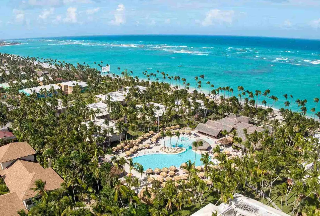 Aerial view of TRS Turquesa and Grand Palladium luxury hotels surrounded by palm trees and turquoise ocean in the Dominican Republic.