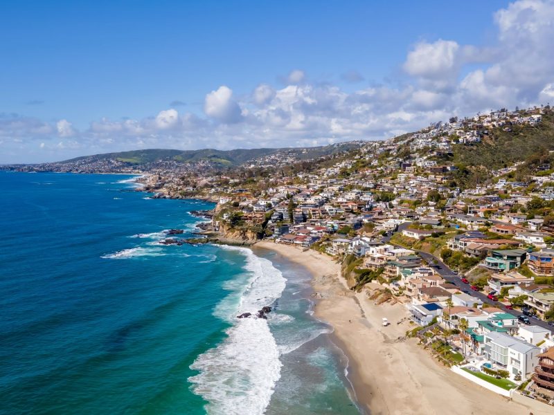 Aerial view of the coastline along Laguna Beach, showcasing sandy beaches, homes, and a vibrant blue ocean under a clear sky.