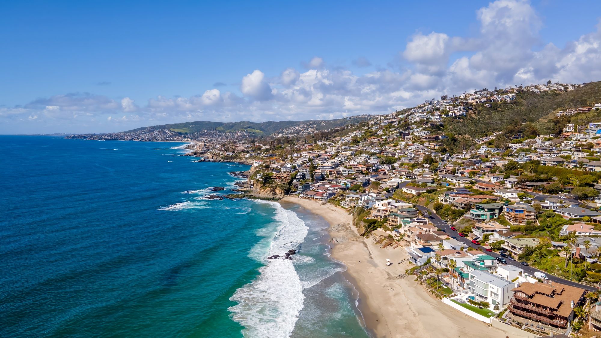 Aerial view of the coastline along Laguna Beach, showcasing sandy beaches, homes, and a vibrant blue ocean under a clear sky.