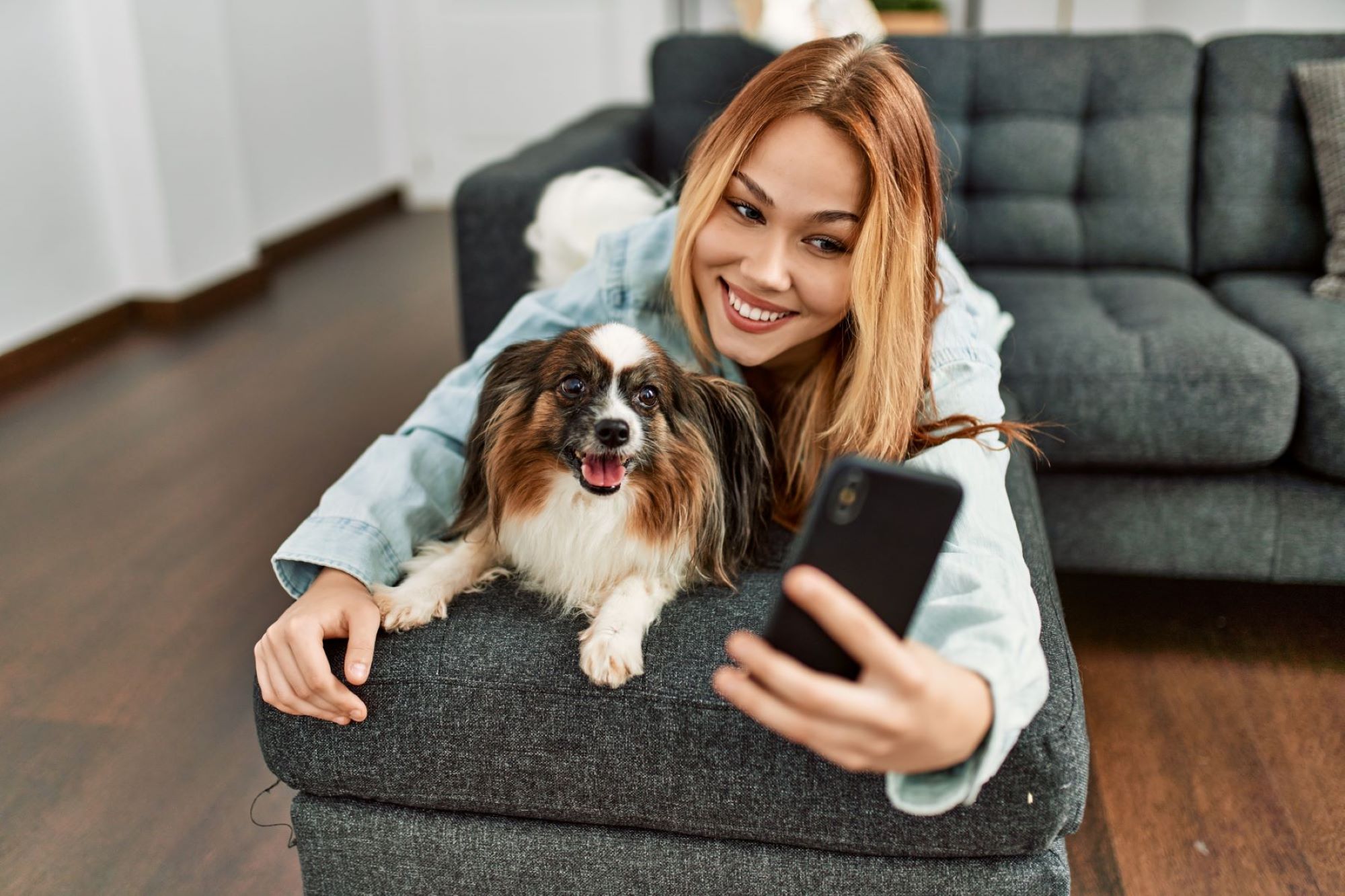 A smiling woman and her dog relax on a couch while she takes a selfie with her smartphone.