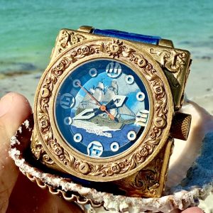 Antique gold watch with intricate details displaying a sailing ship against a blue background, held in a shell by the beach.