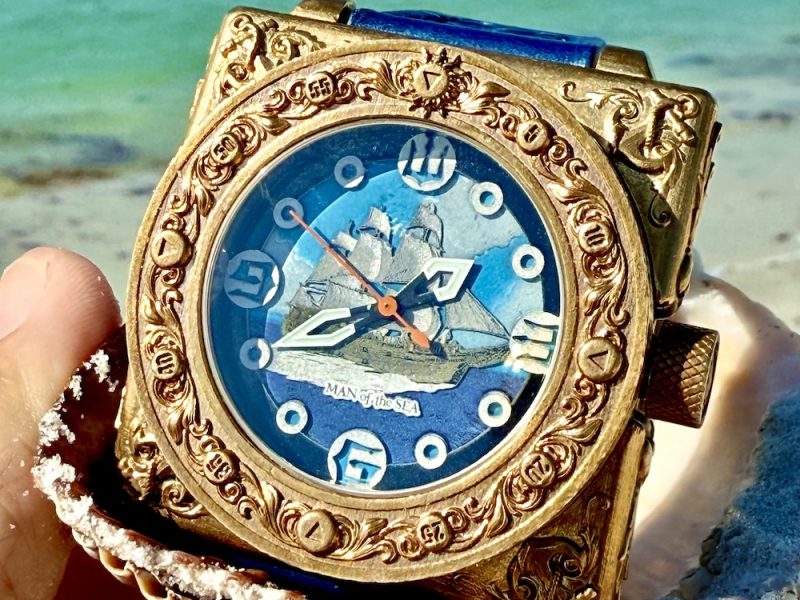 Antique gold watch with intricate details displaying a sailing ship against a blue background, held in a shell by the beach.
