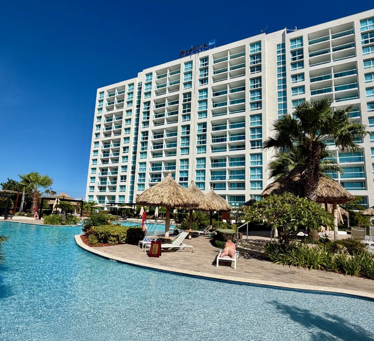 Radisson Blu Aruba features a spacious pool area with tropical plants and cabanas outside the stylish hotel building.