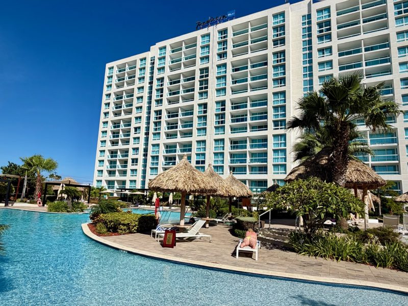 Radisson Blu Aruba features a spacious pool area with tropical plants and cabanas outside the stylish hotel building.
