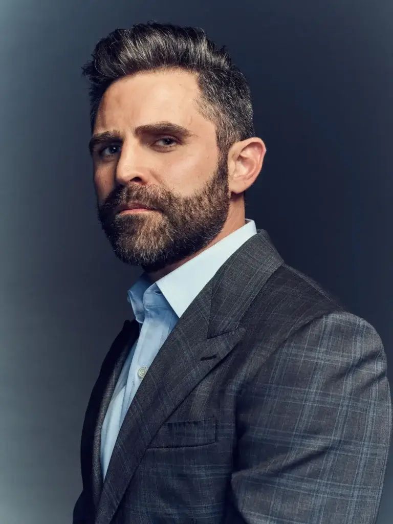 Man with a beard wearing a tailored suit, looking confidently to the side against a dark background.