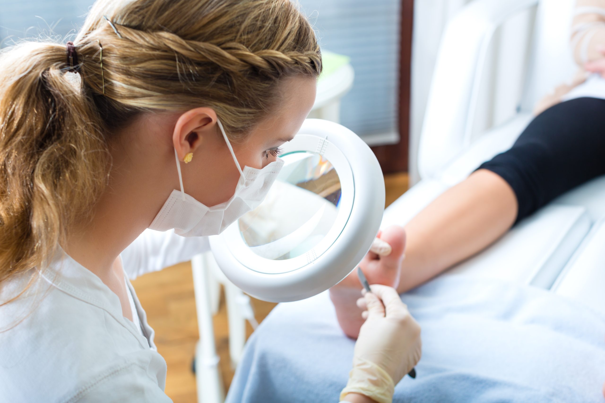 A professional examines a client's foot using a magnifying lamp during a medical pedicure session.