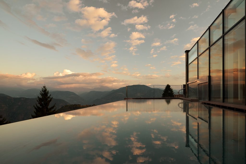 Infinity pool reflecting clouds and mountains at sunset near a modern hotel in the Dolomites.