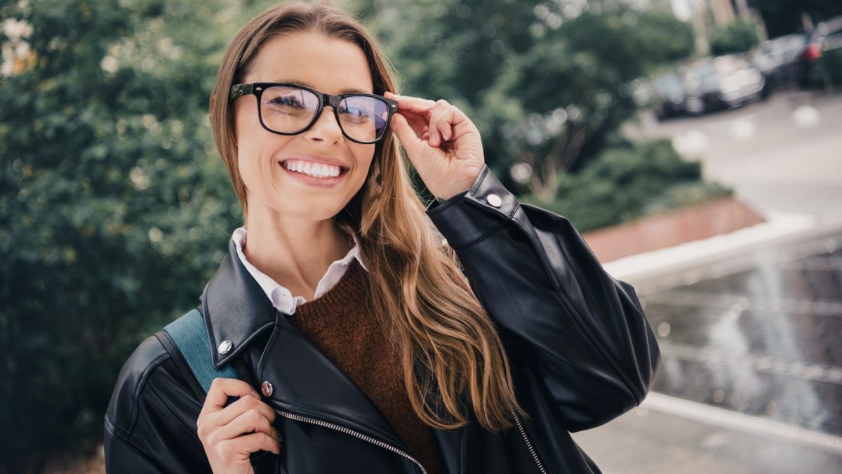 Smiling woman in a leather jacket adjusts her glasses outdoors, showcasing style and confidence in her look.