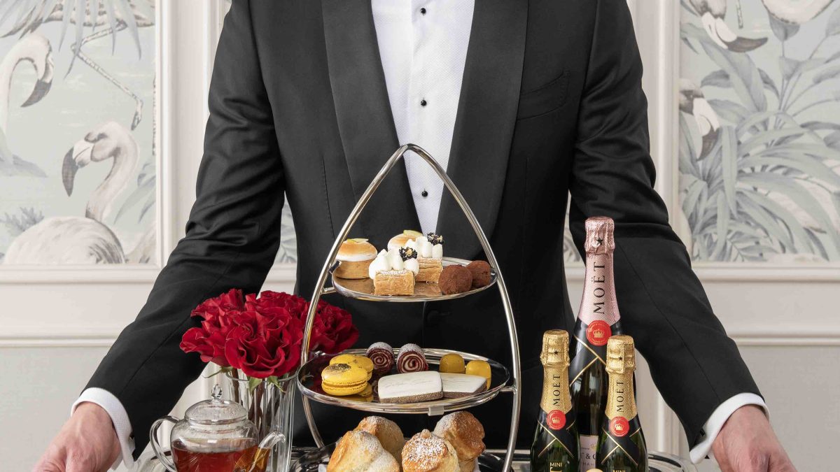 A man in a tuxedo presents an elegant afternoon tea tray featuring scones, pastries, champagne, and detailed garnishes.