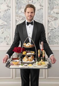 A man in a tuxedo presents an elegant afternoon tea tray featuring scones, pastries, champagne, and detailed garnishes.