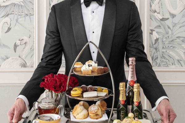 A man in a tuxedo presents an elegant afternoon tea tray featuring scones, pastries, champagne, and detailed garnishes.