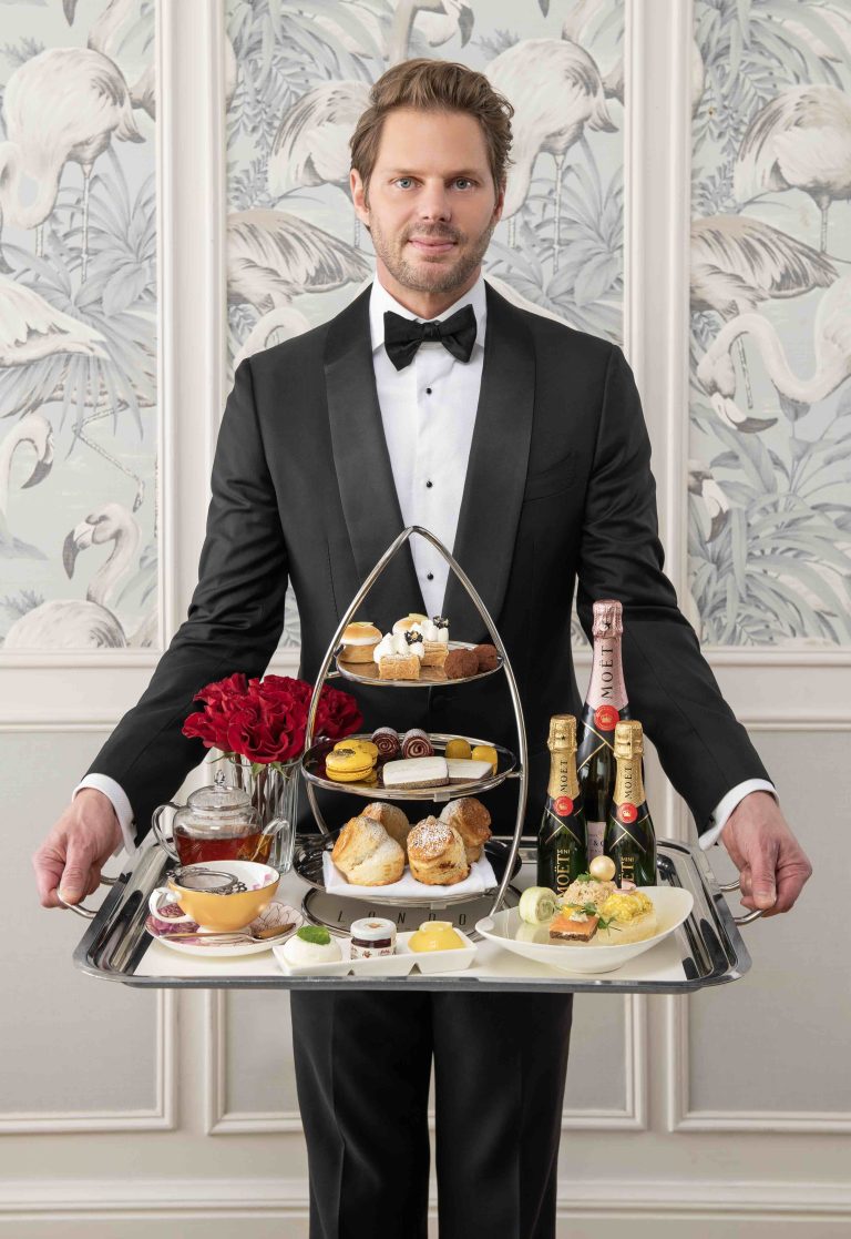 A man in a tuxedo presents an elegant afternoon tea tray featuring scones, pastries, champagne, and detailed garnishes.