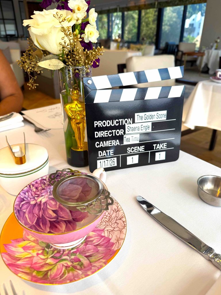 A floral teacup with a silver strainer sits on a decorative saucer next to a film clapboard and a golden trophy, with flowers in a vase.