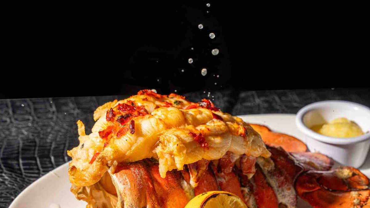 A hand squeezes lemon juice over a lobster tail garnished with herbs, served on a white plate with additional lemon and butter.