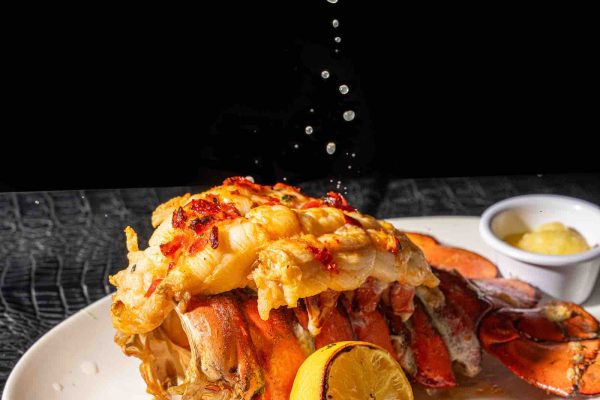 A hand squeezes lemon juice over a lobster tail garnished with herbs, served on a white plate with additional lemon and butter.