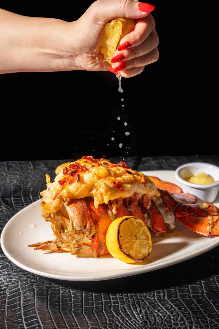 A hand squeezes lemon juice over a lobster tail garnished with herbs, served on a white plate with additional lemon and butter.