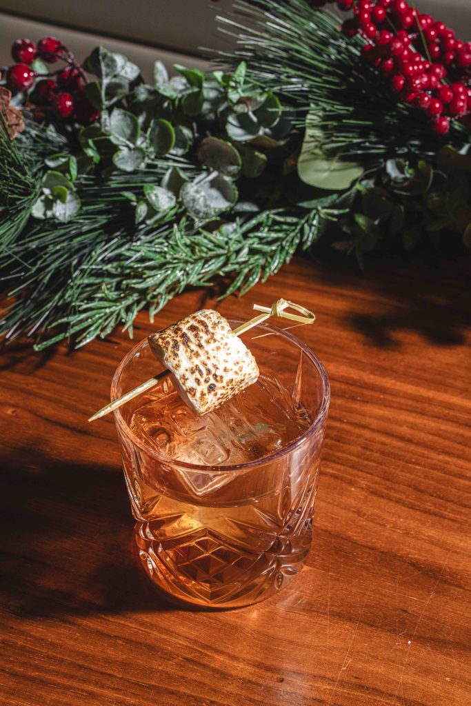 A glass of cocktail with a toasted marshmallow garnish, surrounded by festive greenery and berries.