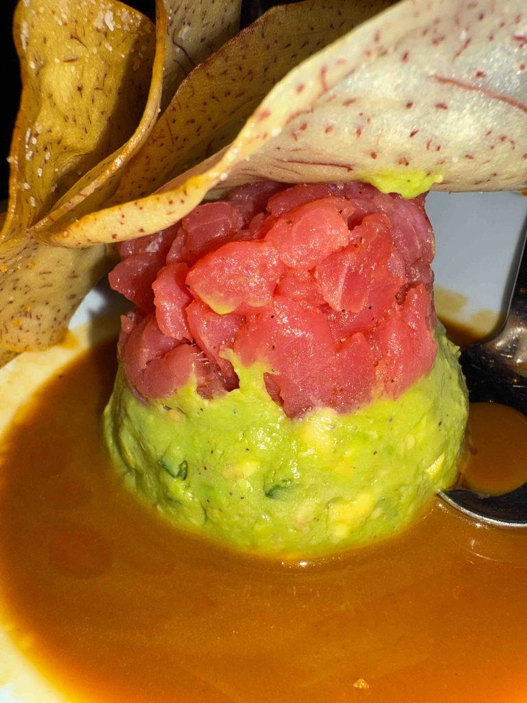 A serving of ahi tuna tartare perched on avocado mousse, accompanied by a crispy garnish and drizzled with sauce.