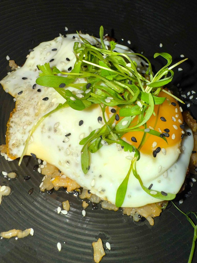 Fried egg topped with green sprouts and sesame seeds on a bed of rice in a black bowl.