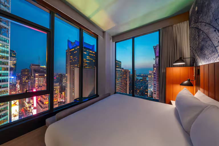 Modern bedroom with a city view at Tempo by Hilton, showcasing skyscrapers and vibrant lights at dusk.