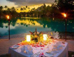 A beautifully set dinner table adorned with flowers, candles, and a view of a tranquil lagoon at sunset.