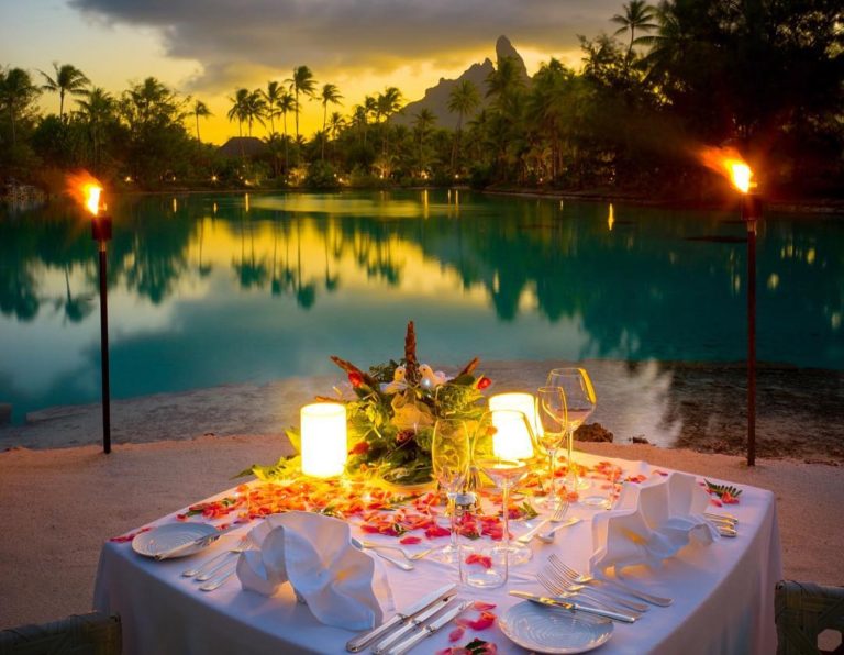 A beautifully set dinner table adorned with flowers, candles, and a view of a tranquil lagoon at sunset.